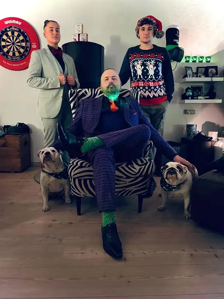 Three people and two bulldogs pose in a living room. The central man sits in a zebra-print chair wearing a plaid suit, green…