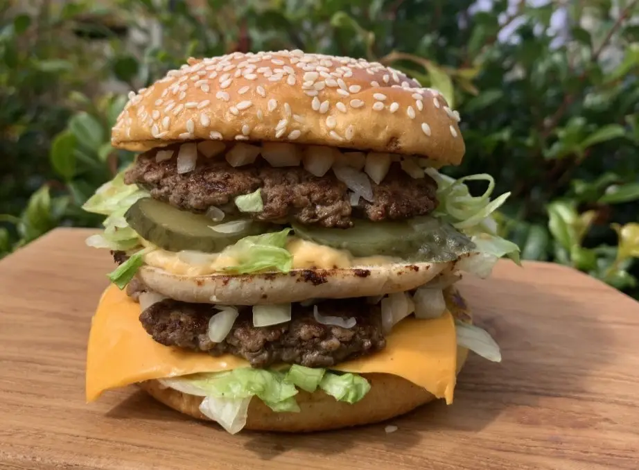 a large, double-patty cheeseburger with pickles and onions sits on a wooden board, topped with a sesame seed bun, against a…