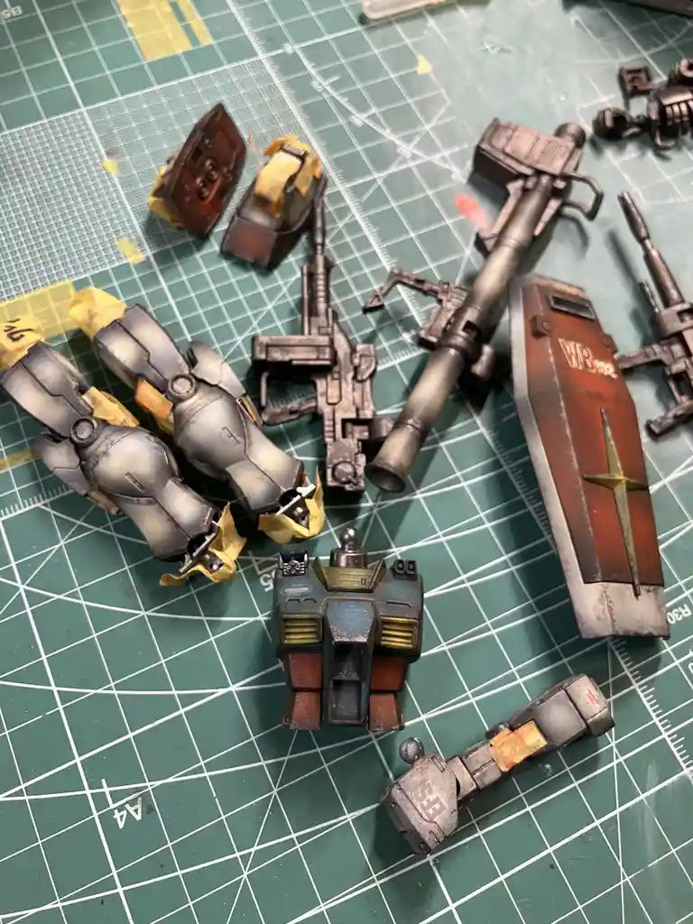 A collection of painted and weathered mecha model parts, including arms, weapons, and a shield, are scattered on a green…
