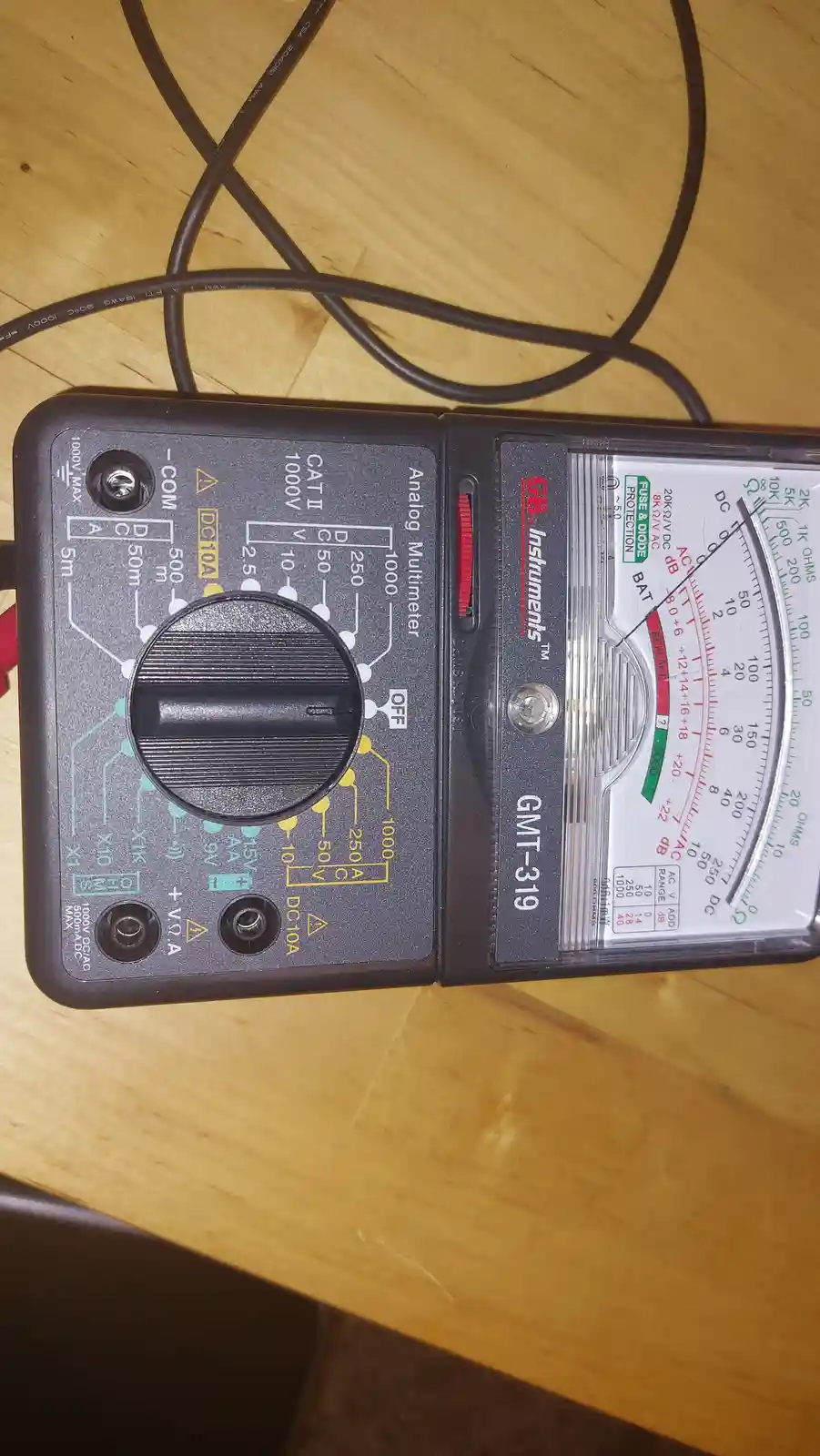 An analog multimeter, model GMT-319 by GB Instruments, sits on a wooden surface with its needle pointing to zero. The device…