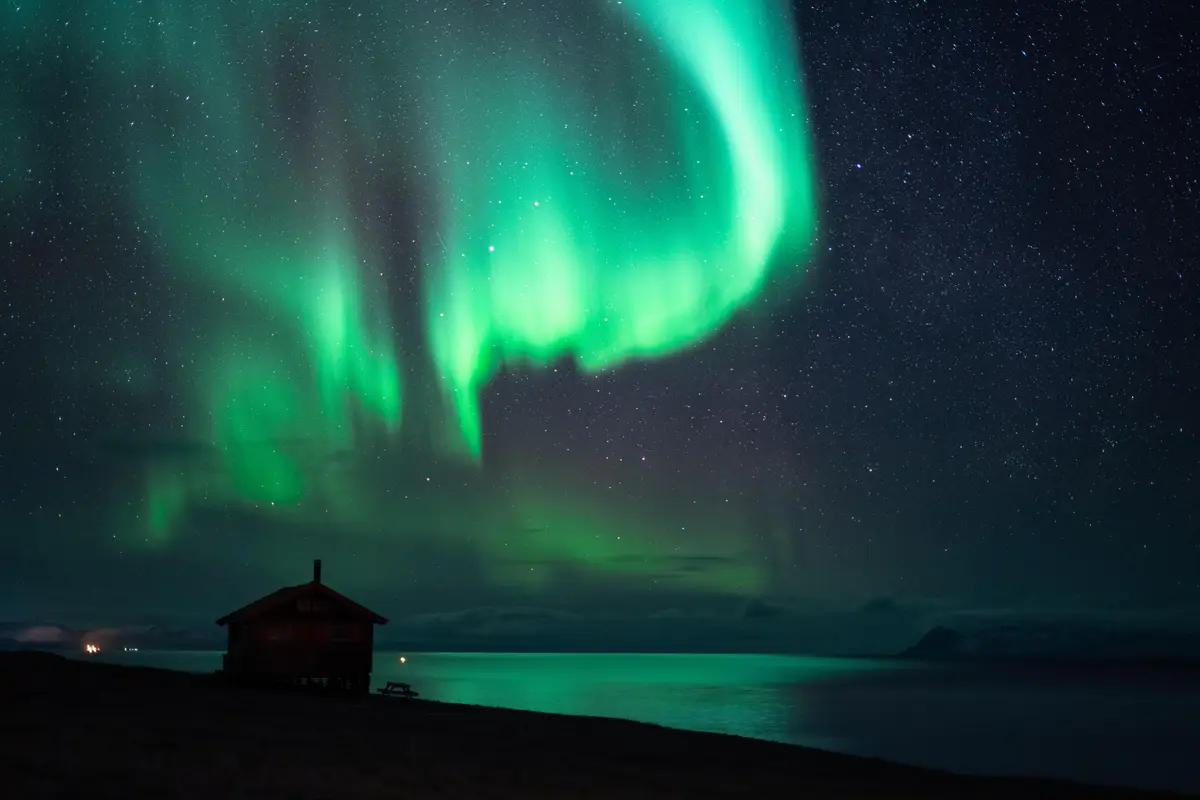 A serene night scene shows a small wooden cabin on a shoreline under a starry sky, with vibrant green auroras dancing…