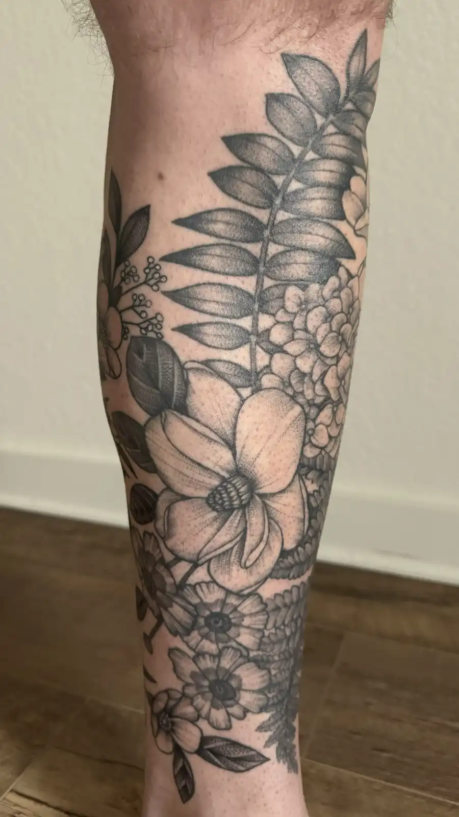 A close-up photo shows a person's forearm and lower arm covered in a detailed black-and-gray floral tattoo featuring large…