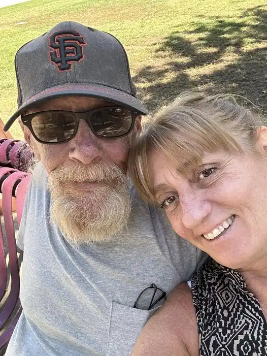 A man with a white beard and sunglasses wearing a San Francisco Giants baseball cap sits next to a smiling woman in a…