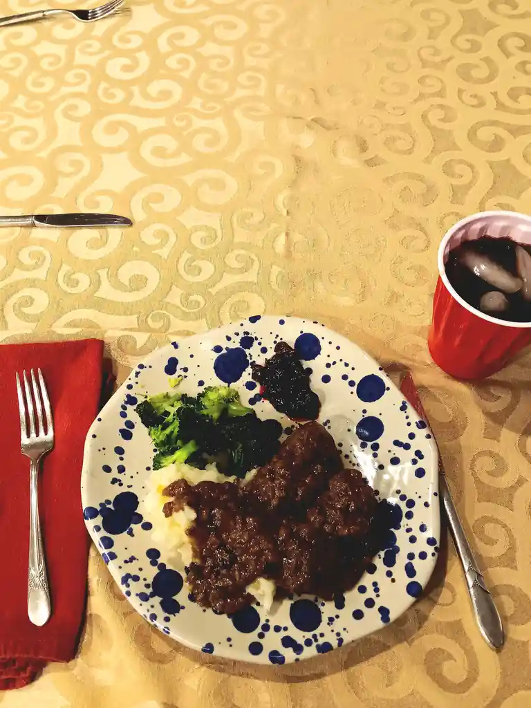 A plate of meatloaf with gravy, mashed potatoes, and broccoli sits on a patterned tablecloth next to a red cup of iced soda…
