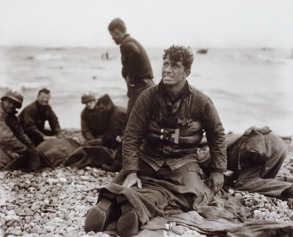 A black-and-white historical photo shows a weary, smiling soldier sitting on a pebbled beach, wearing a life vest and…