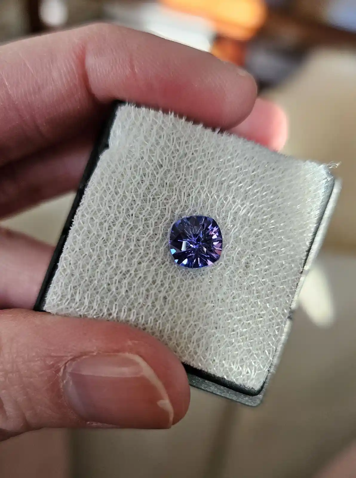 A hand holds a small, square, white cushioned jewelry display with a round, faceted, deep purple-blue gemstone centered on…