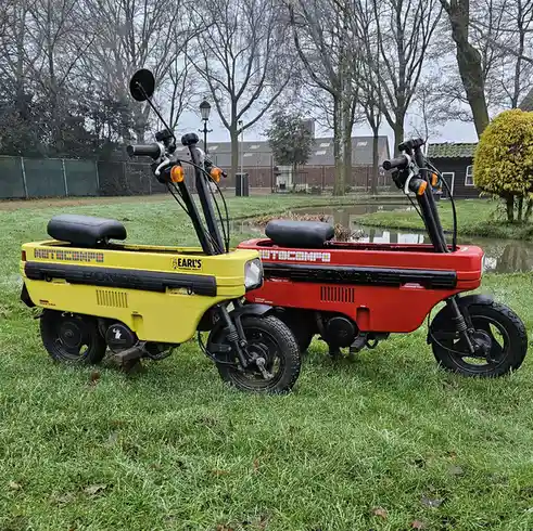 Two vintage Honda Motocompo three-wheeled scooters, one yellow and one red, parked side-by-side on grass near a pond. Both…