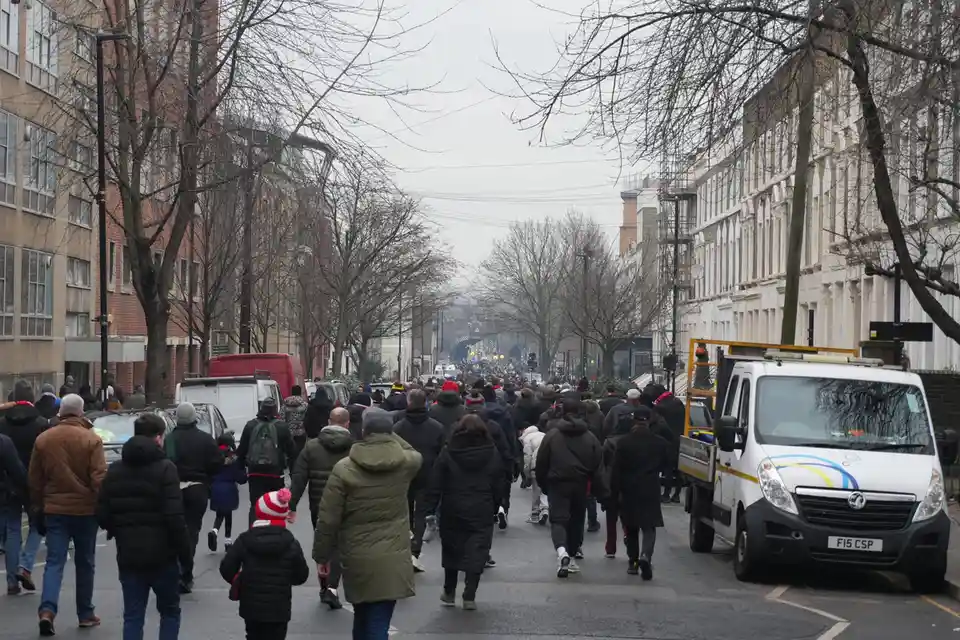 A wide street is filled with a dense crowd of people walking away from the camera toward a distant archway, many wearing…