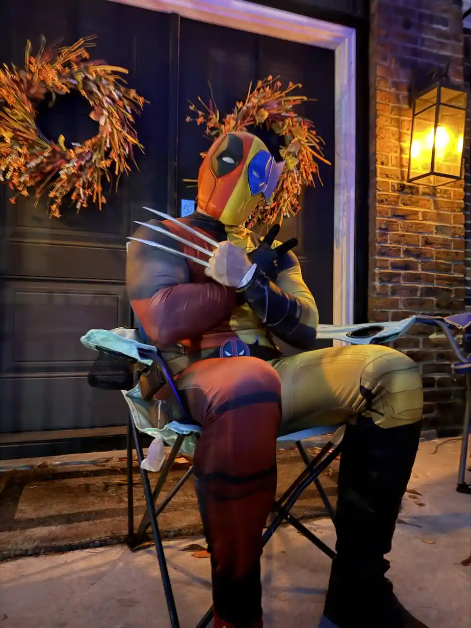 A person in a Deadpool costume sits in a folding chair outside a house, with two autumn wreaths placed on their head like…