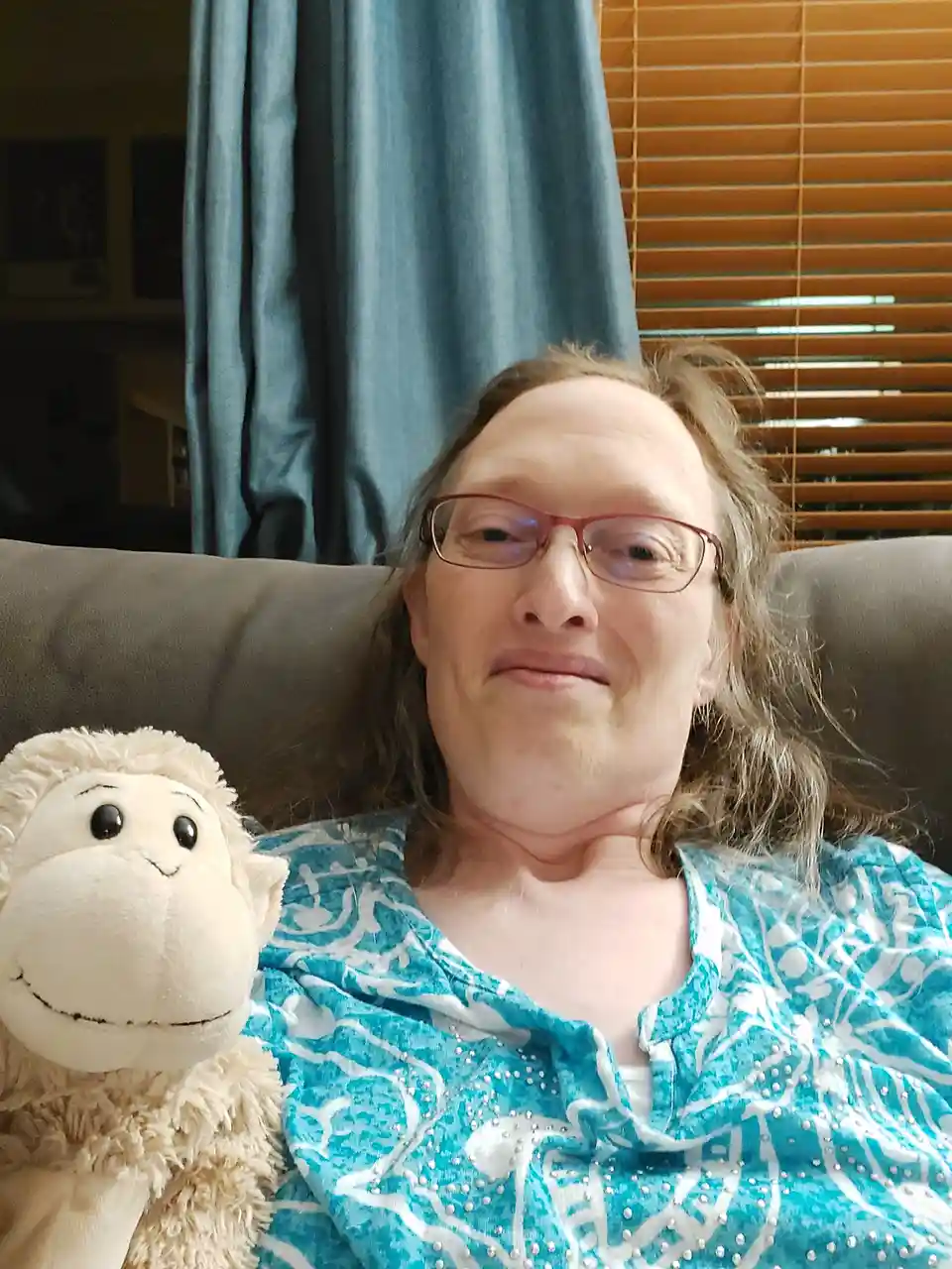 A person with glasses and long hair sits on a couch next to a white stuffed monkey, both looking at the camera with a…