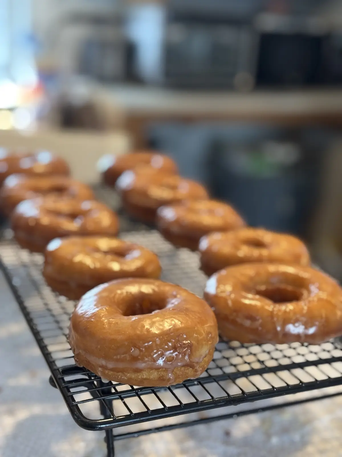 A close-up photo shows rows of freshly glazed, golden-brown donuts cooling on a black wire rack. The donuts are glossy with…