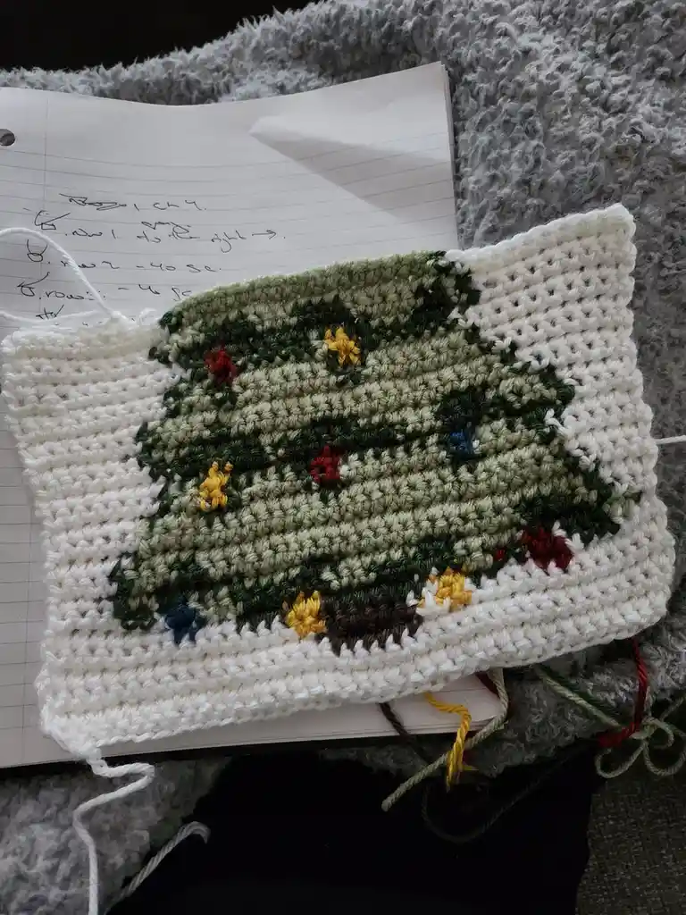 A close-up photo shows a partially completed crocheted Christmas tree with green, white, and multicolored yarn, resting on a…