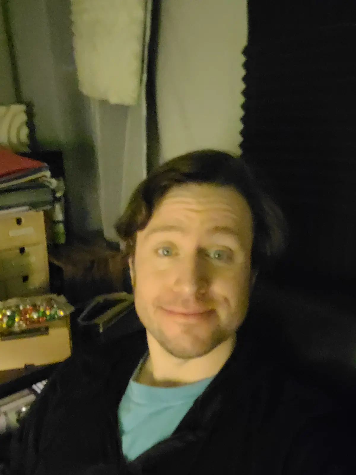 A blurry selfie of a man with a smug, slightly unsettling smile, wearing a black jacket over a teal shirt, in a dimly lit…