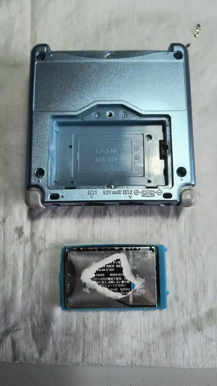 The image shows the back of a silver handheld gaming device with its battery compartment open, revealing a blue plastic…