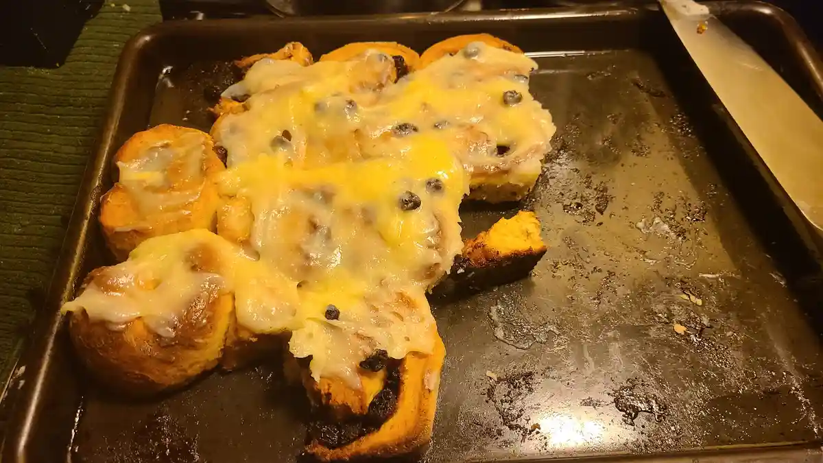 A baking sheet holds partially eaten cinnamon rolls covered in melted cheese and raisins, with one roll missing and the rest…