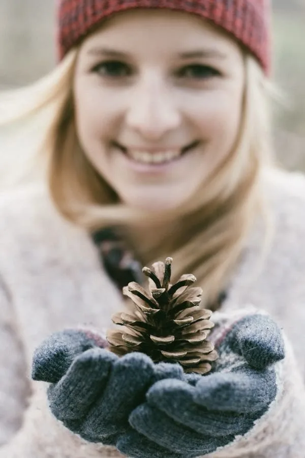 A smiling woman wearing a red plaid beanie and blue knit gloves holds a pine cone in her cupped hands. The image is softly…