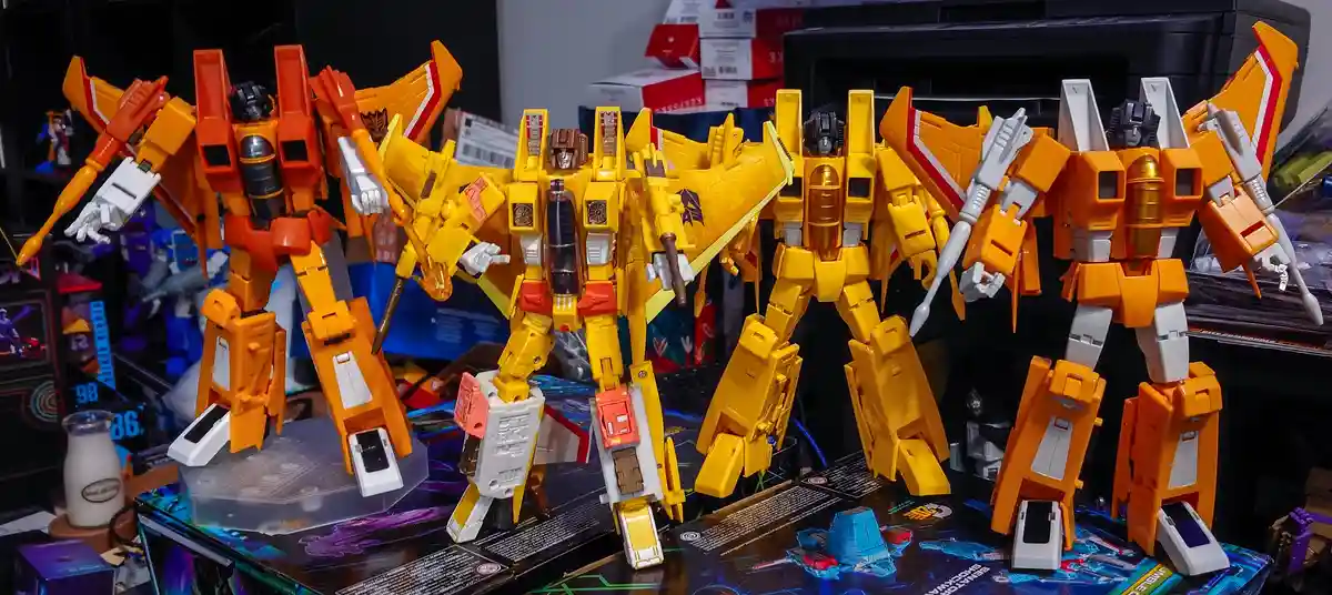 Five large, detailed Transformers action figures, mostly yellow and orange, are displayed on a cluttered desk with boxes and…