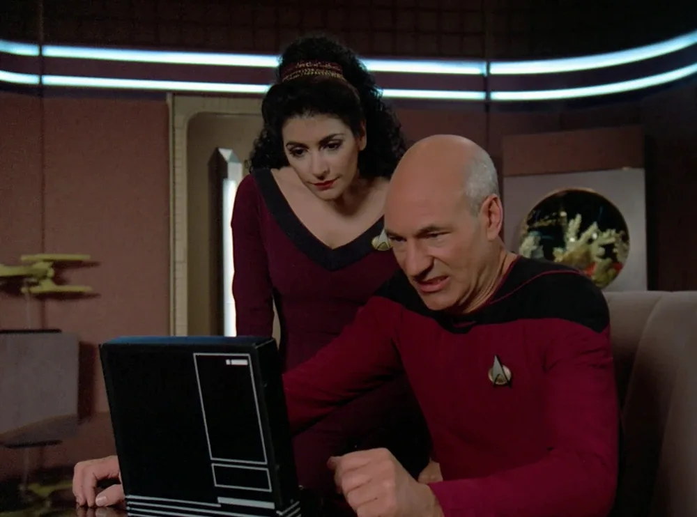 A man in a red Starfleet uniform sits at a desk with a black laptop, looking frustrated while a woman in a matching uniform…