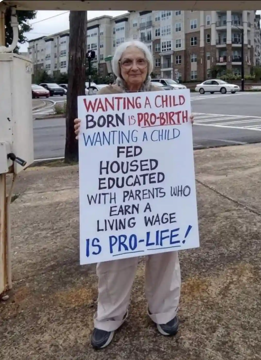 An elderly woman with white hair and glasses stands outdoors holding a handmade white sign with red and blue handwritten…