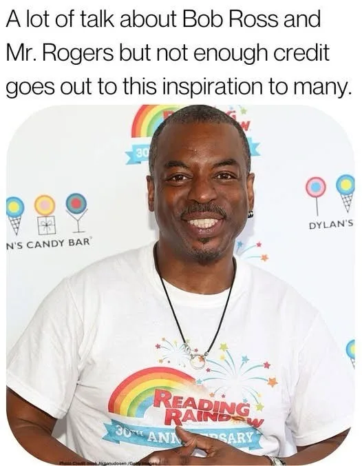 A smiling Black man with short hair and a goatee, wearing a white t-shirt with a rainbow and fireworks graphic that reads…