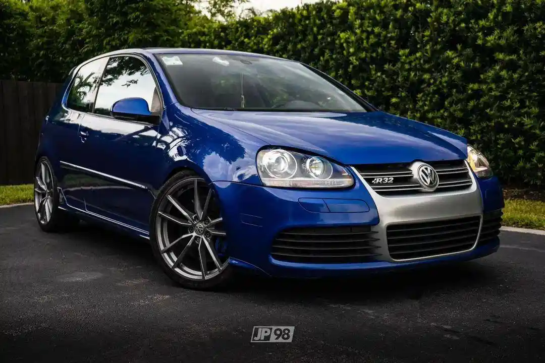 A glossy blue Volkswagen Golf R32 is parked on asphalt with green hedges in the background. The car has a sporty front…