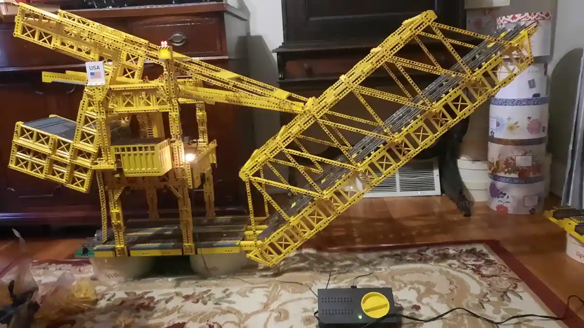 A large, intricate yellow Lego model of a construction crane is displayed indoors on a patterned rug, leaning precariously…