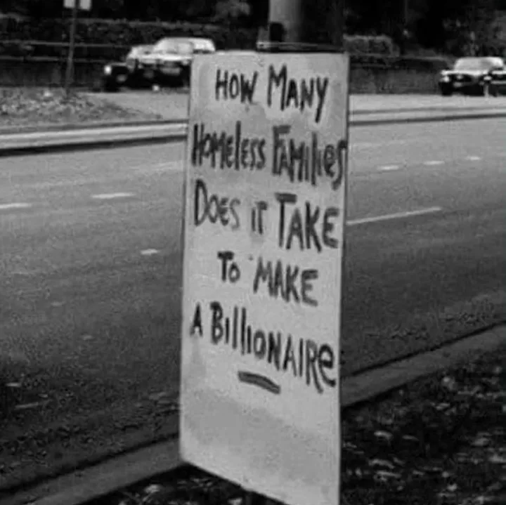 A black-and-white photo shows a handwritten sign on a roadside, asking 'HOW MANY HOMELESS FAMILIES DOES IT TAKE TO MAKE A…