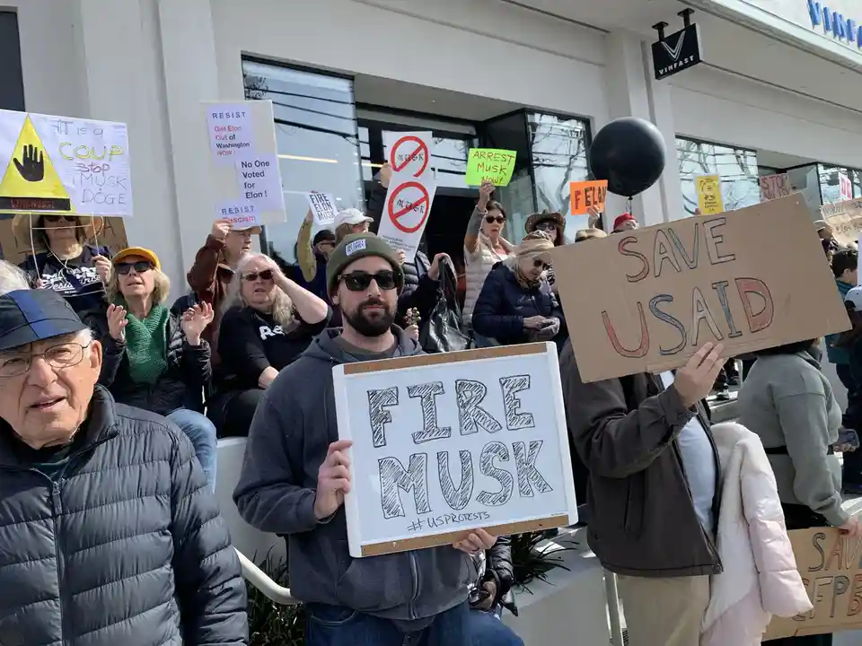 A crowd of protesters holds various signs outside a VinFast building, including one that says 'FIRE MUSK' on a whiteboard…
