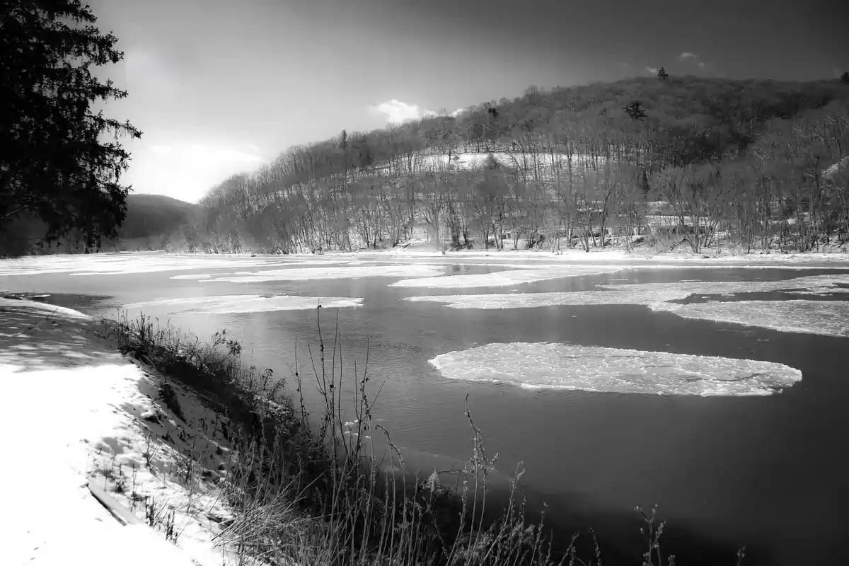 A black-and-white photo of a partially frozen river in winter, with snow-covered banks and bare trees on hillsides. Ice…