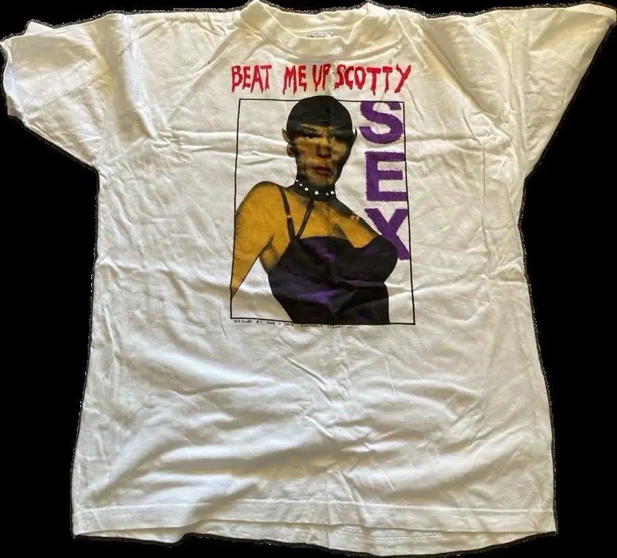 A white t-shirt displays a graphic of a woman in a purple top with the text 'BEAT ME UP SCOTTY' in red above her and the…