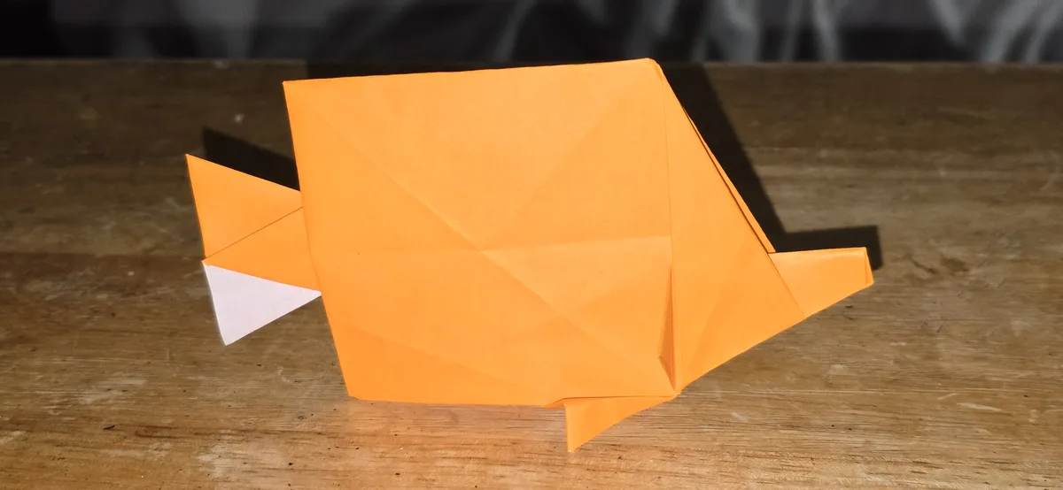 An orange origami fish with a white triangular tail fin sits on a wooden table. The fish has a simple, geometric design with…