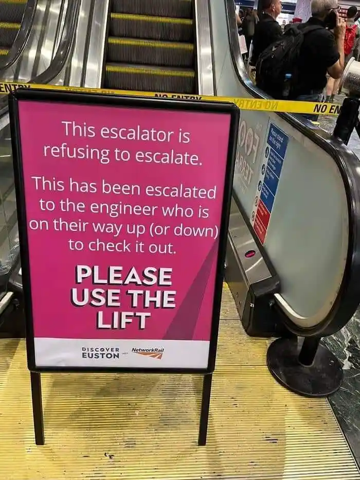 A pink sign stands in front of a broken escalator, with yellow 'NO ENTRY' tape above it. The sign humorously states that the…