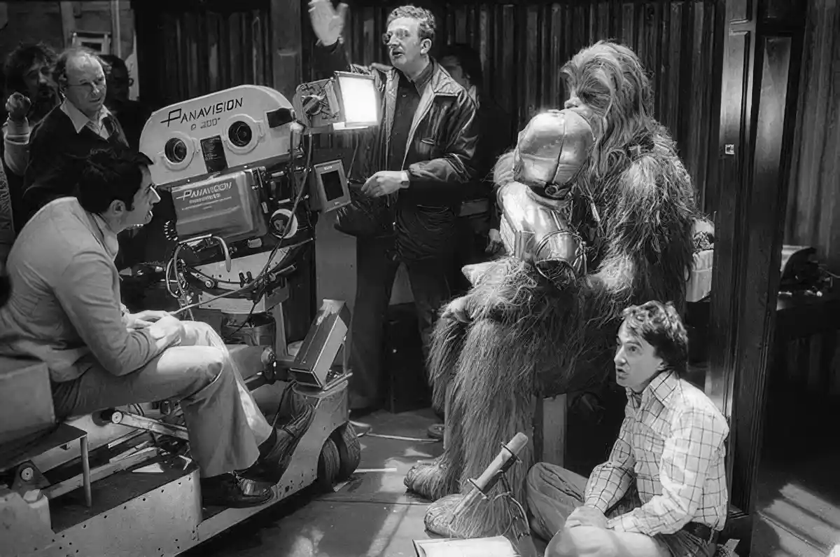 A black-and-white behind-the-scenes photo from a Star Wars film set shows director George Lucas gesturing near a Panavision…