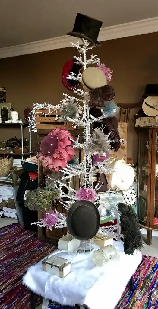 A white, sparkly, wire-frame Christmas tree is decorated with various hats, including top hats, bowler hats, and feathered…