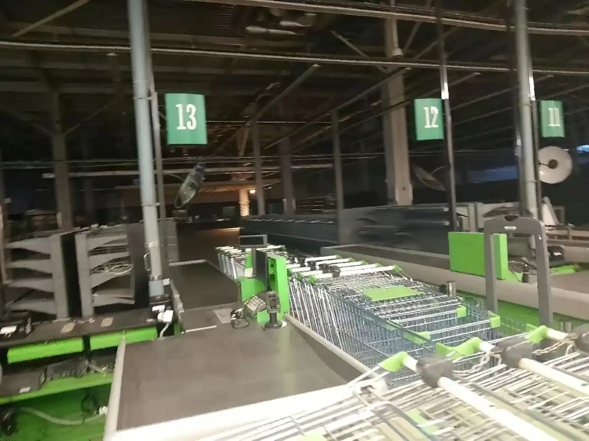 An empty supermarket checkout area with rows of unattended shopping carts and numbered green signs hanging from the ceiling…