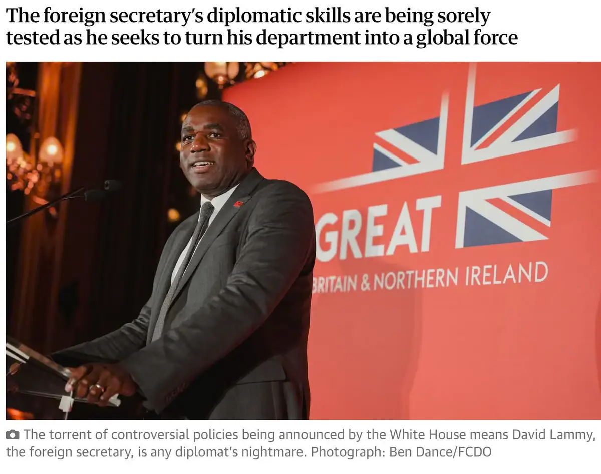 A man in a dark suit and tie stands at a podium in front of a red backdrop featuring a large Union Jack and the word 'GREAT'…