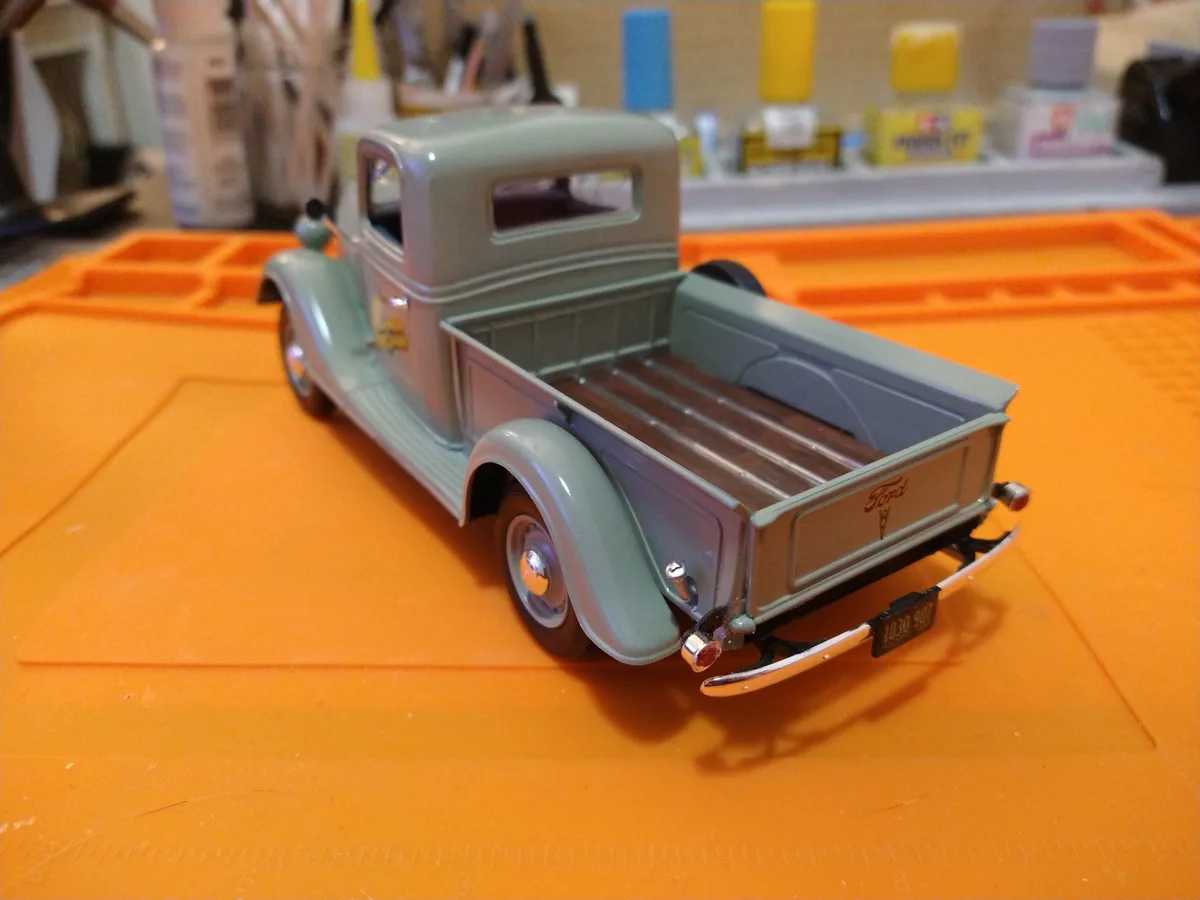 A light green vintage Ford pickup truck model sits on an orange work mat, with a workshop background showing paint supplies…