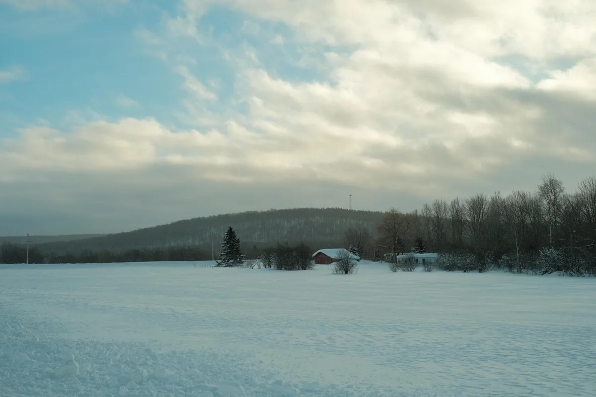 A wide snowy landscape with a red barn and a white trailer near a forested hill under a cloudy sky. The overlaid text reads…