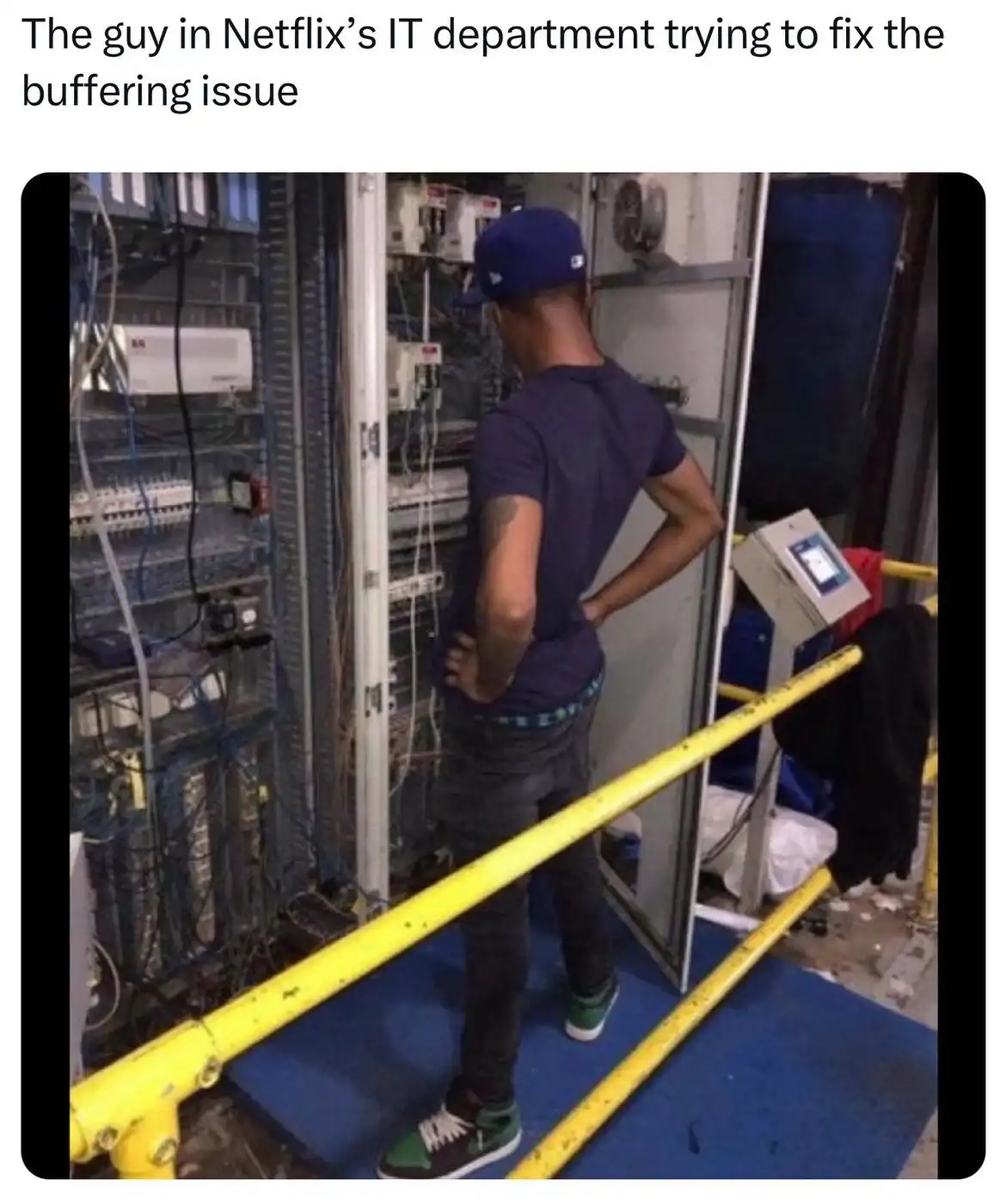 A man in a blue cap and t-shirt stands with his hands on his hips in front of a large server rack filled with cables and…