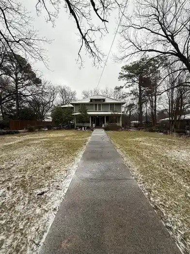 A photo shows a residential house viewed from a paved walkway, with light snow scattered across the grassy lawn under bare…