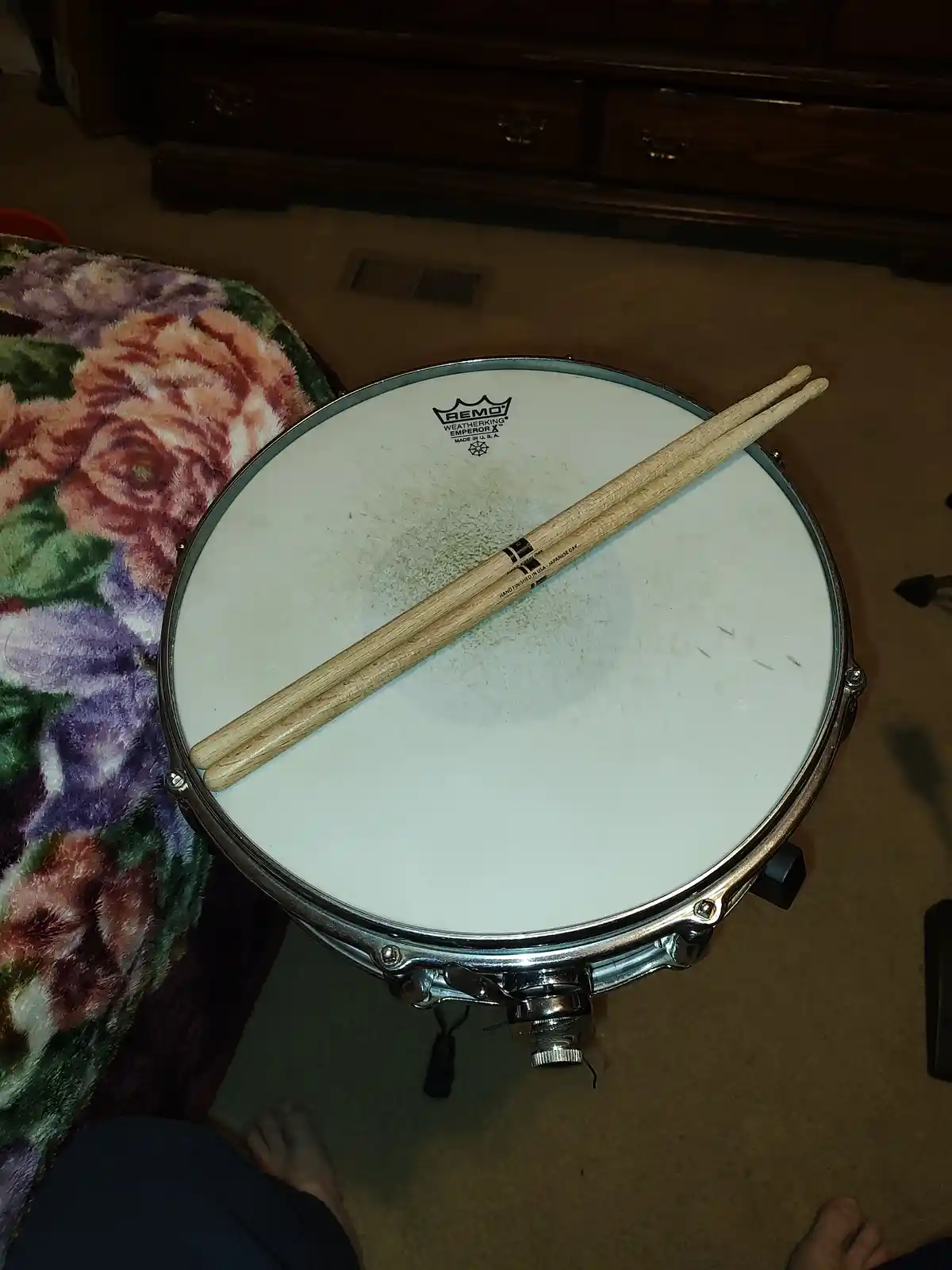 A close-up photo shows a snare drum with two drumsticks resting diagonally across its white drumhead. The drum has a chrome…