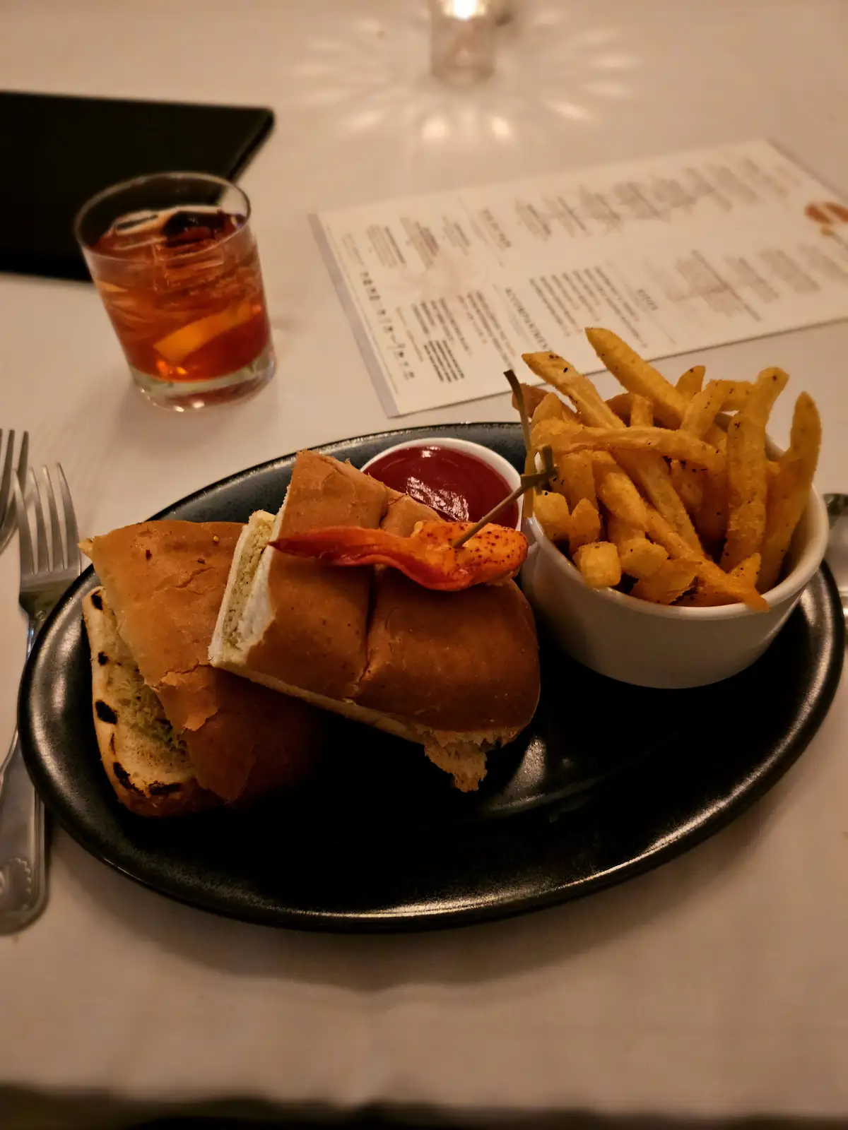 A close-up photo of a meal on a dark plate, featuring a sandwich cut in half, a small bowl of fries, and a side of ketchup…