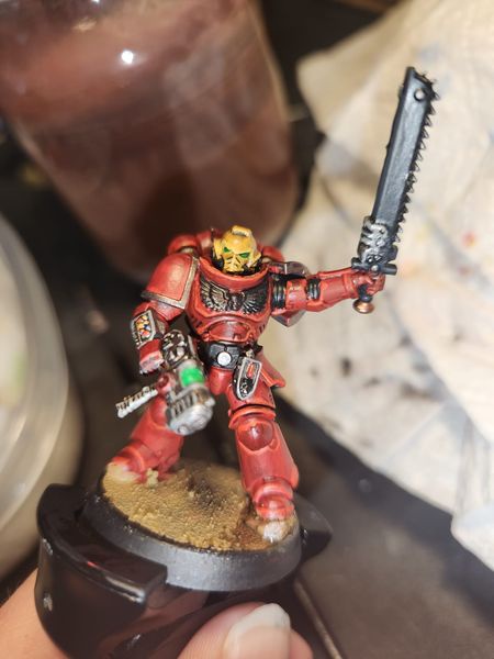 A hand holds a painted miniature of a red-armored warrior with a chainsword, standing on a circular base. The figure has a…