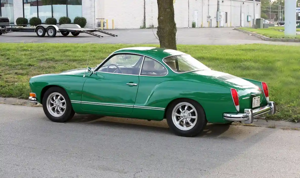 A green vintage Karmann Ghia coupe is parked on an asphalt road next to a grassy area with a tree. The car has chrome…