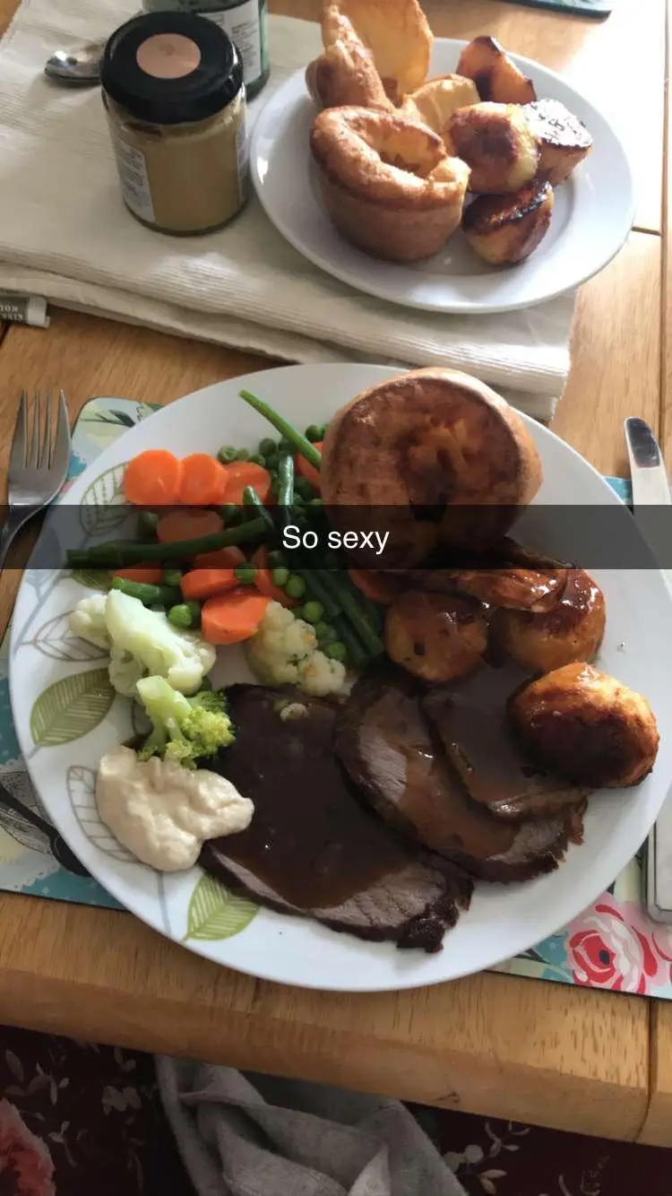 A close-up photo of a full English roast dinner on a white plate, featuring sliced roast beef with gravy, roasted potatoes…