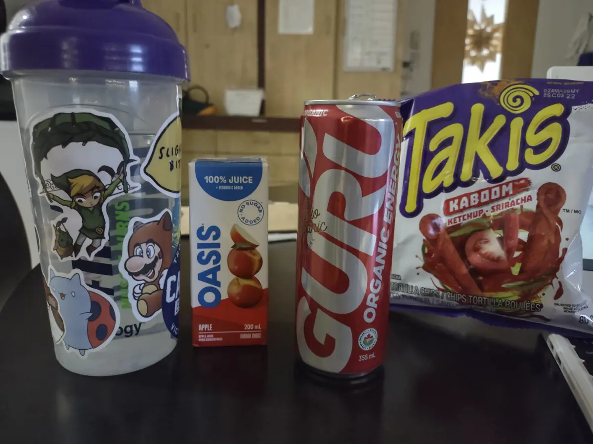 A clear plastic shaker cup with cartoon stickers, a small box of apple juice, a can of Guri organic energy drink, and a bag…