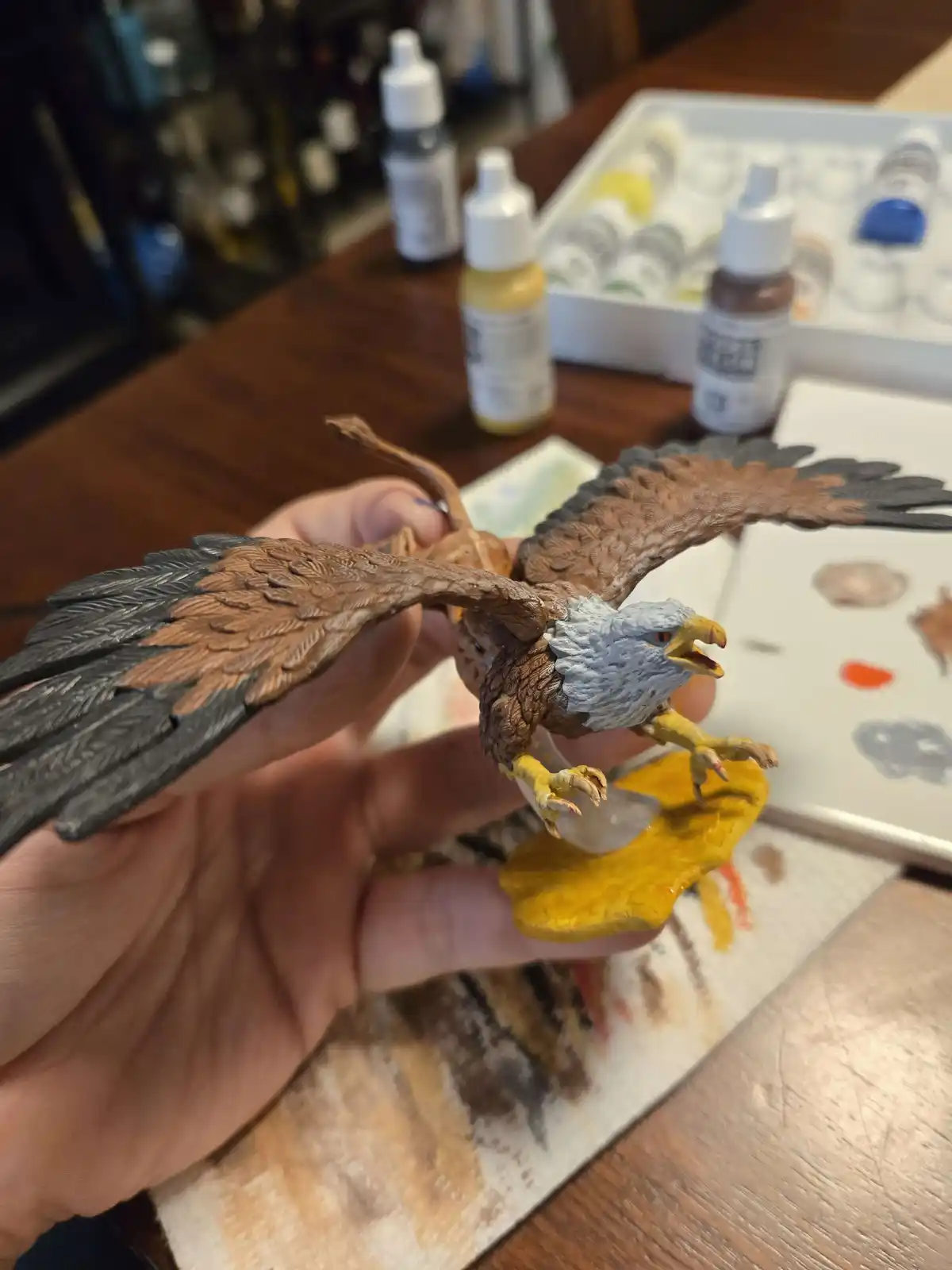 A hand holds a painted eagle figurine with brown wings, white head, and yellow beak and talons, standing on a yellow base…