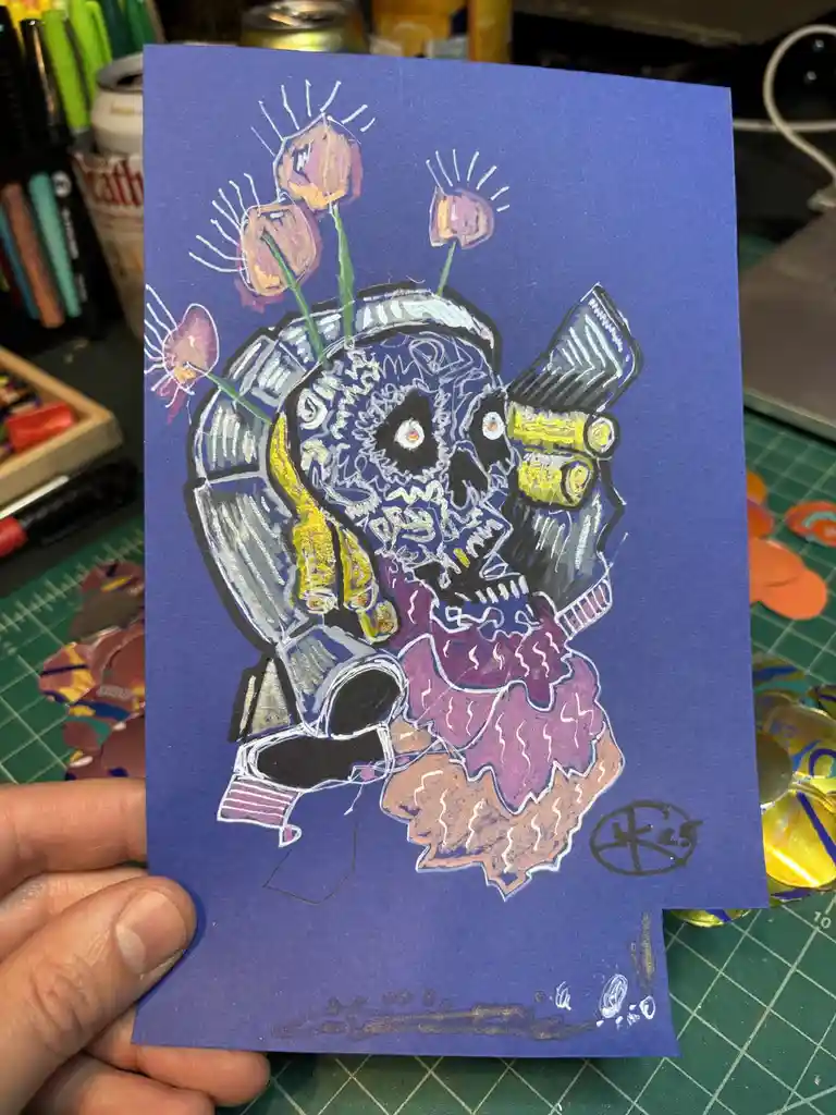 A hand holds a blue paper drawing of a stylized skull wearing a helmet and goggles, adorned with purple flowers and a purple…