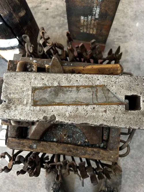 A close-up photo shows a heavily worn, rusty metal block with a rectangular cutout and a square hole, sitting atop a pile of…