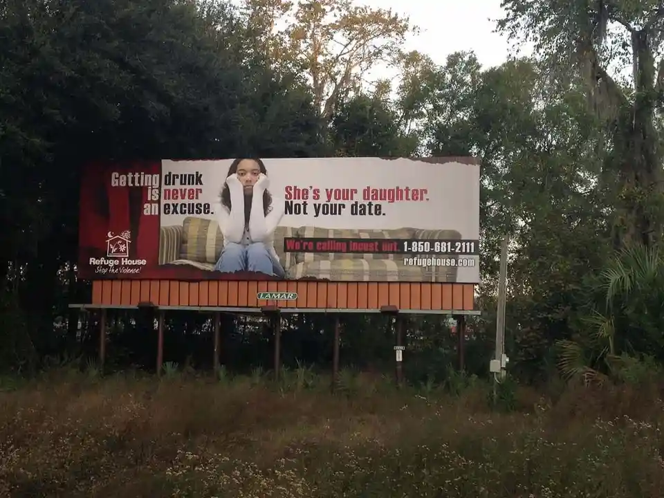 A large outdoor billboard features a photo of a young woman sitting on a couch with her hands covering her face, conveying…
